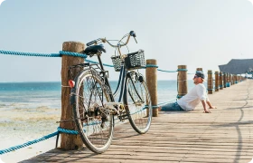 Мужчина наблюдает за океаном, сидя на причале, рядом стоит велосипед с корзиной.