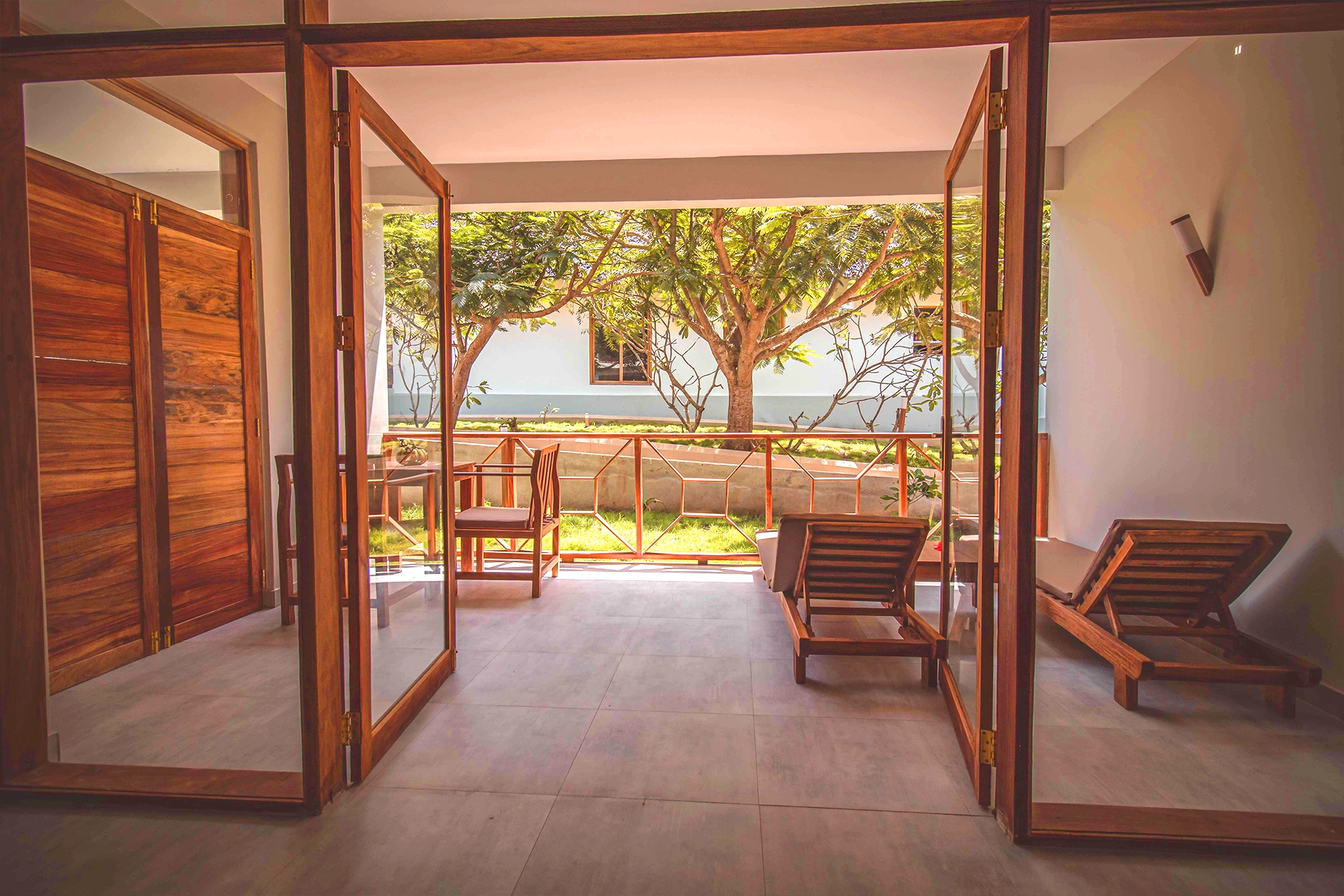 Widok z pokoju hotelowego na taras wyposażony w leżaki i stół z krzesłami, z egzotyczną, zanzibarską przyrodą w tle.