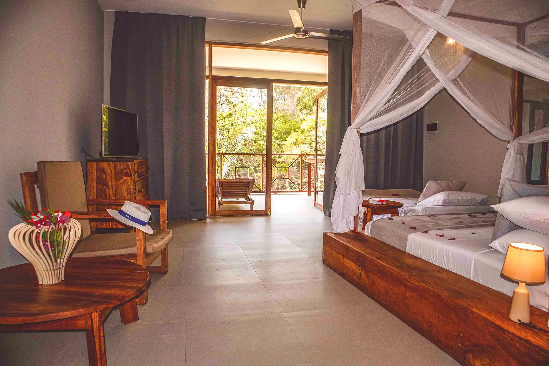 Elegancki pokój, wyposażony w drewniane meble – dwa łóżka, stolik, fotel i szafka pod telewizor, z wyjściem na balkon i widokiem na egzotyczną przyrodę.